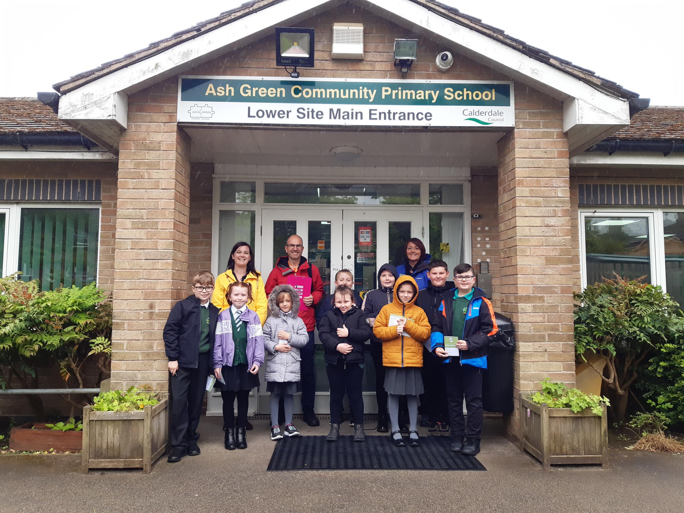 A group of adults and children sand outside the entrance of a school. The signs reads 'Ash Green Community Primary School lower Site Main Entrance'