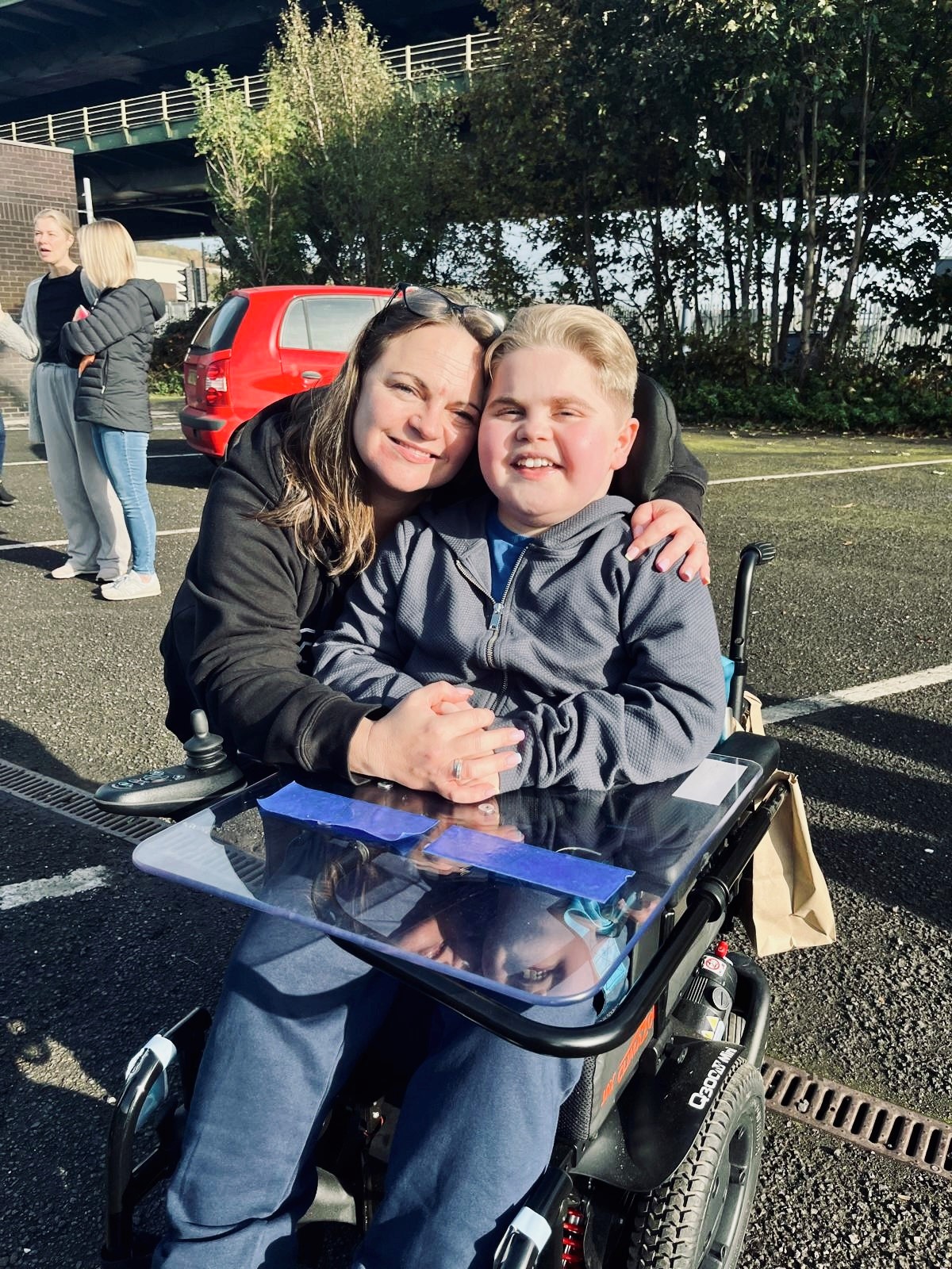a woman holds her son close to her, he is in a power wheelchair. They are holding hands and smiling.