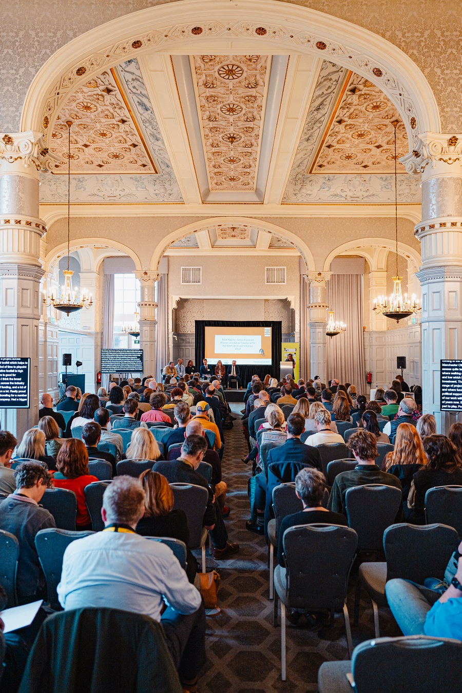 A hotel conference room is filled with people. They are facing toward a presentation on screen.