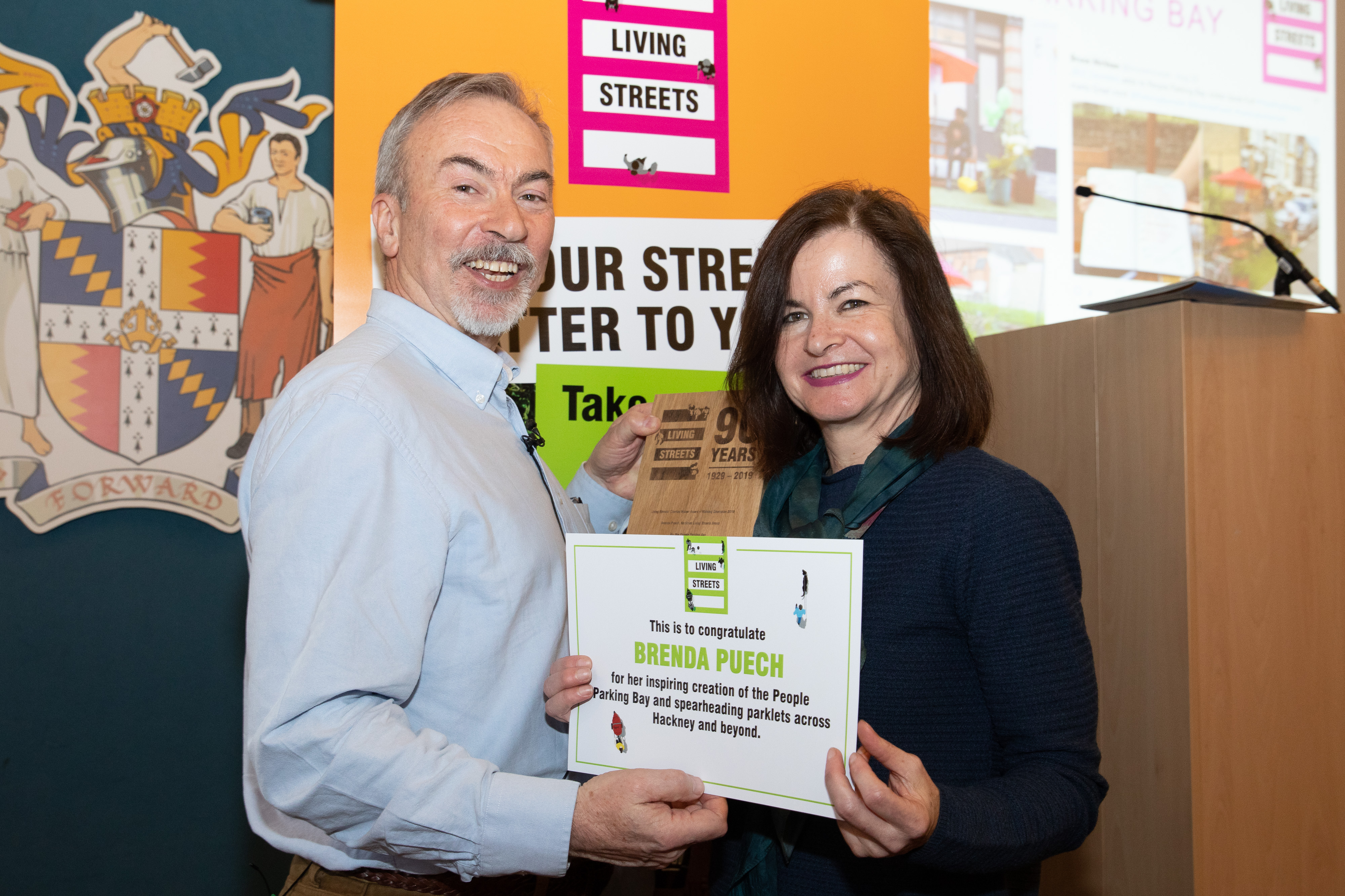 Brenda Puech is standing next to a man and they are both holding a certificate with her name on