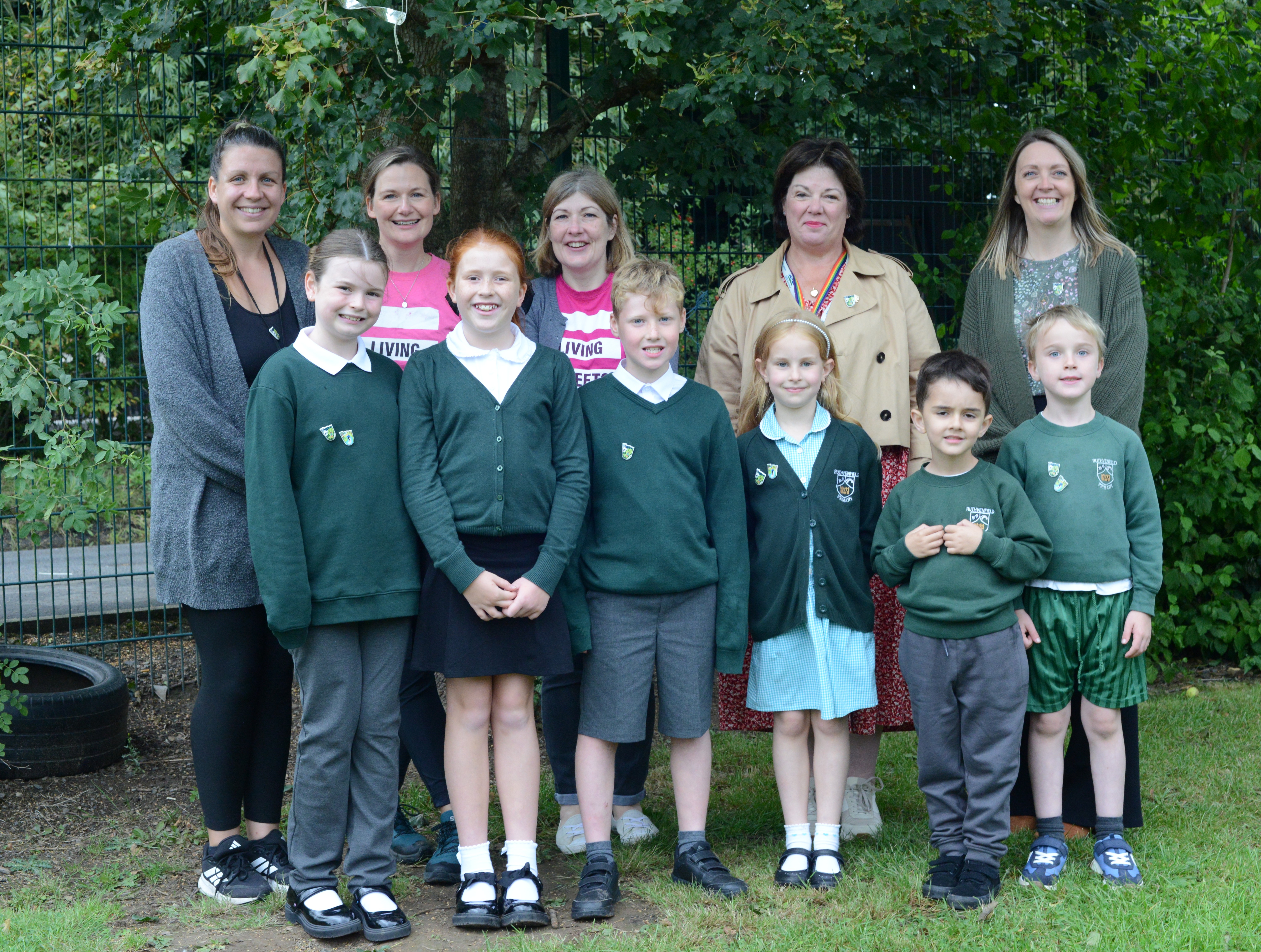 A group of school children and adults are standing and smiling together in front of some trees