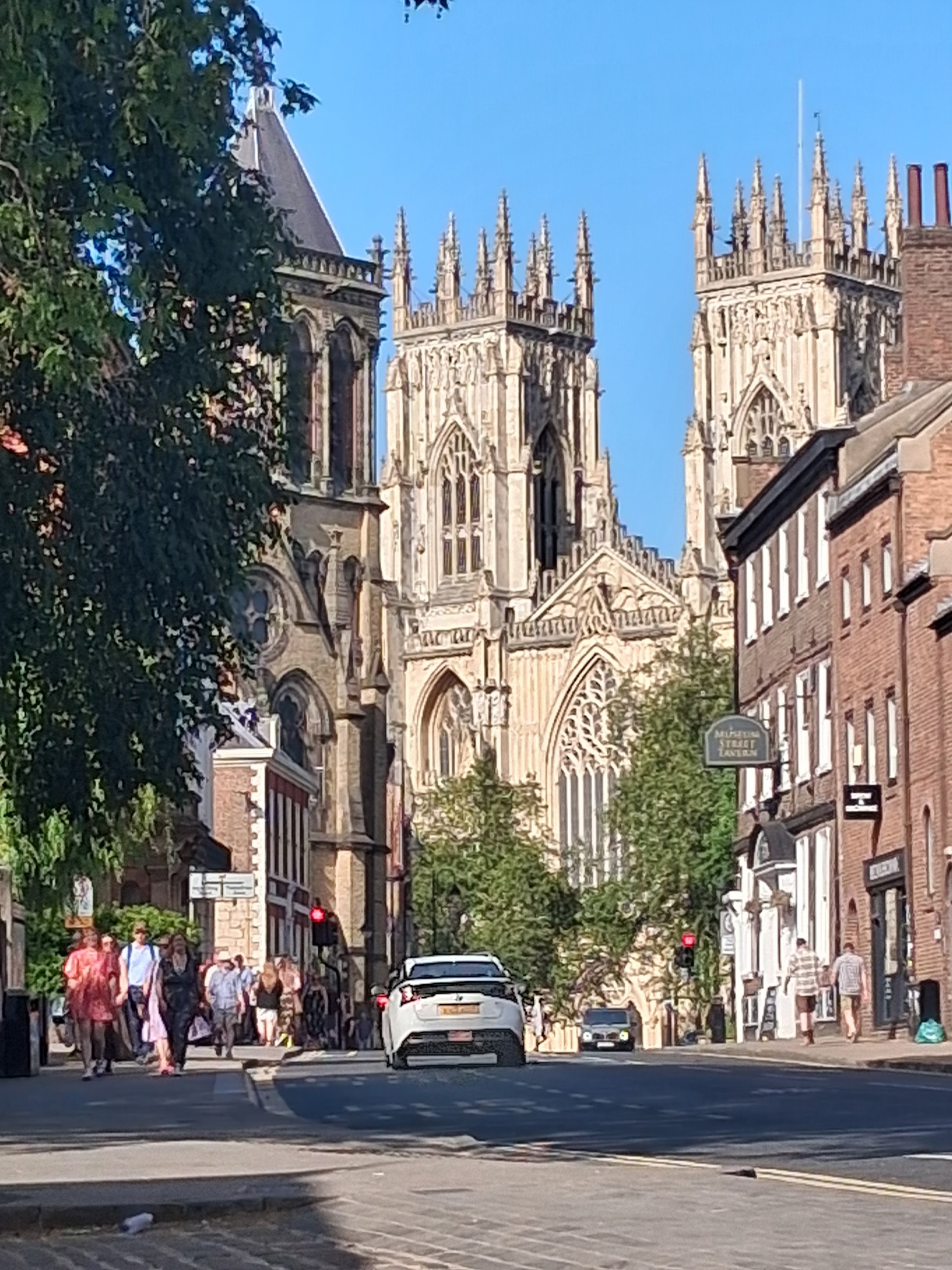 A photograph of a tree lined street leading up to York Minster. The sky overhead is blue and cloudless.