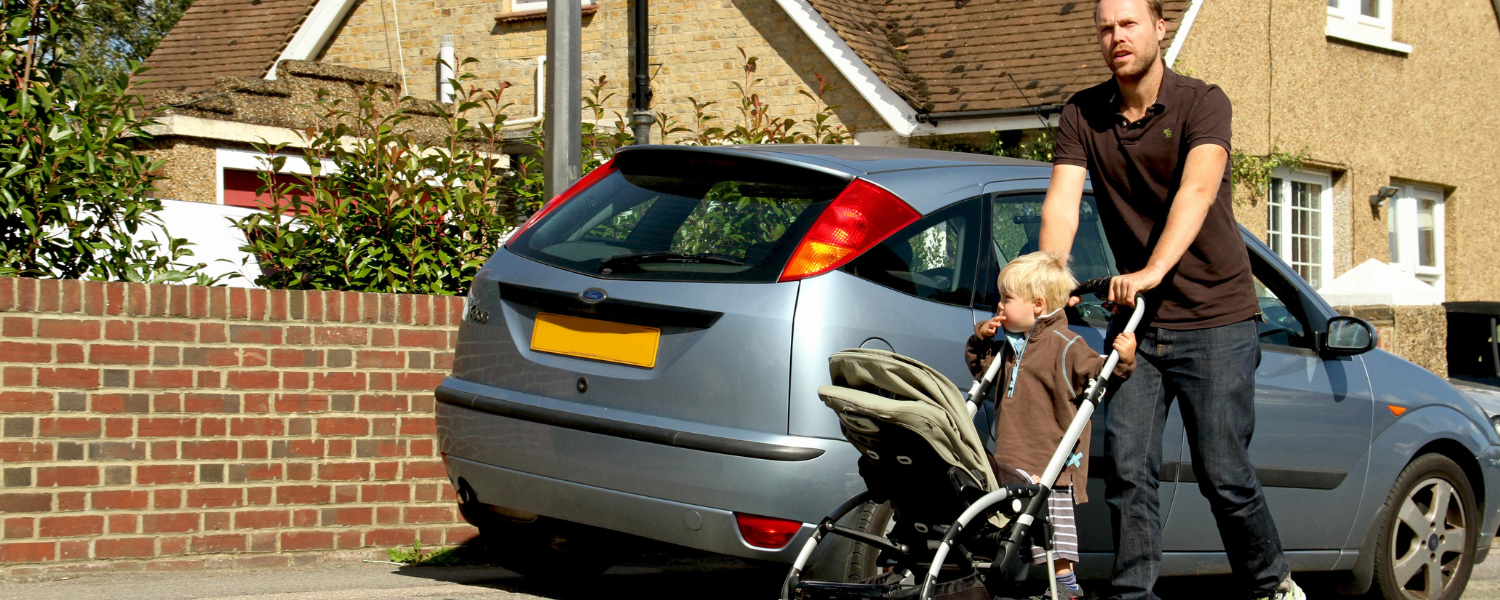 man struggles to push buggy around car parked on pavement