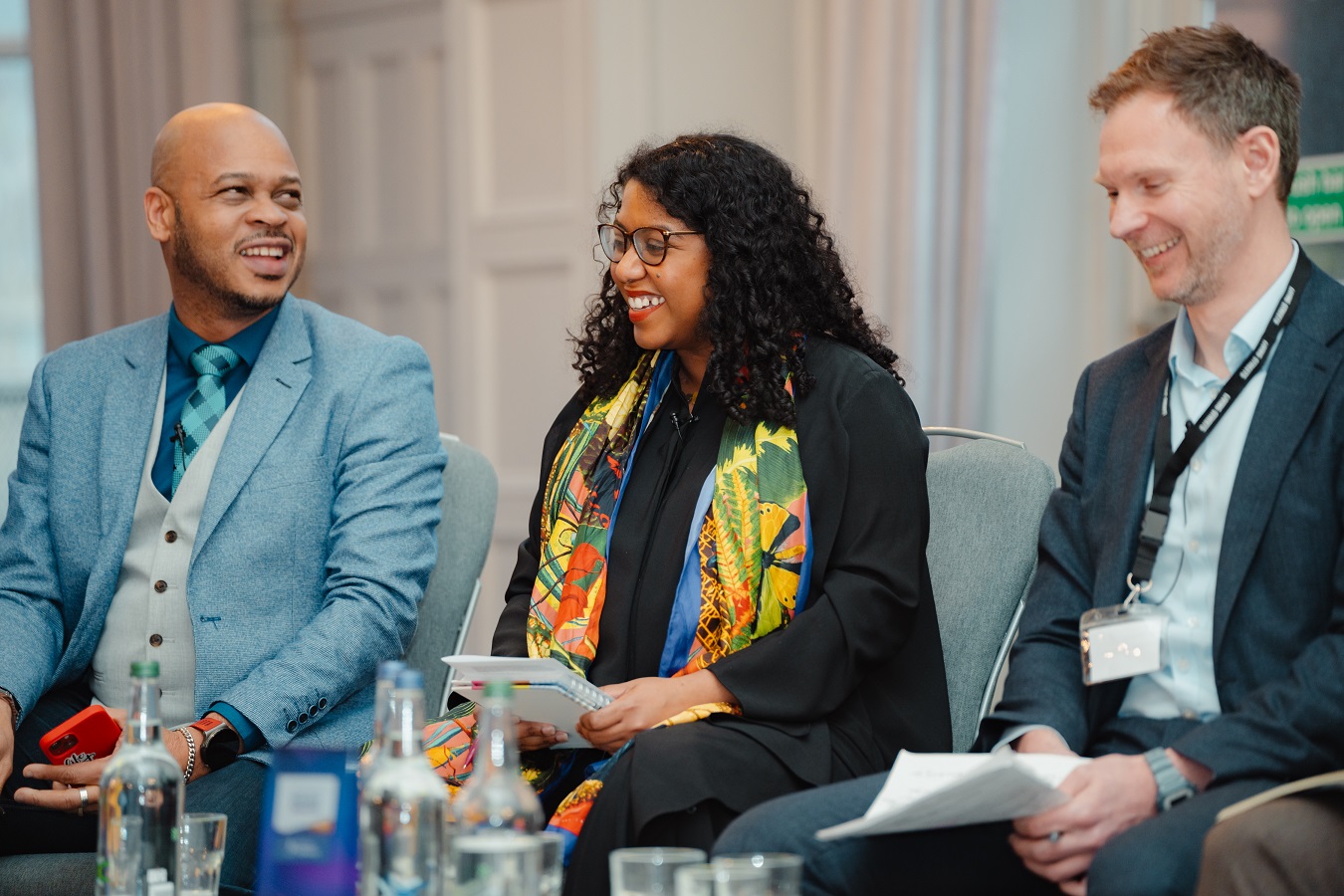 Three people are presenting on a stage, they are smiling mid-conversation