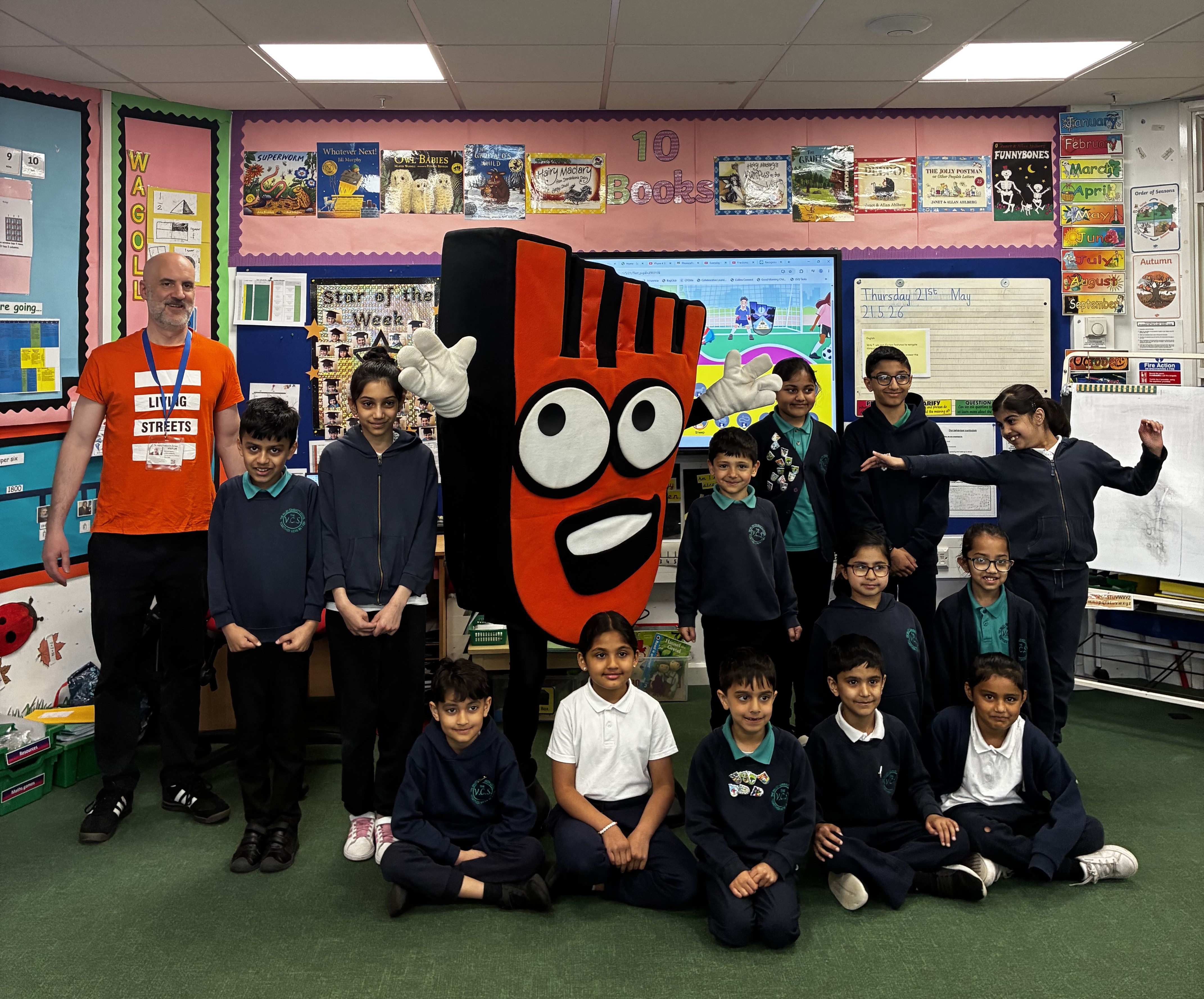 An image of school children wearing blue and green uniforms alongside an adult wearing an orange Living Streets tshirt and Strider, Living Streets' mascot