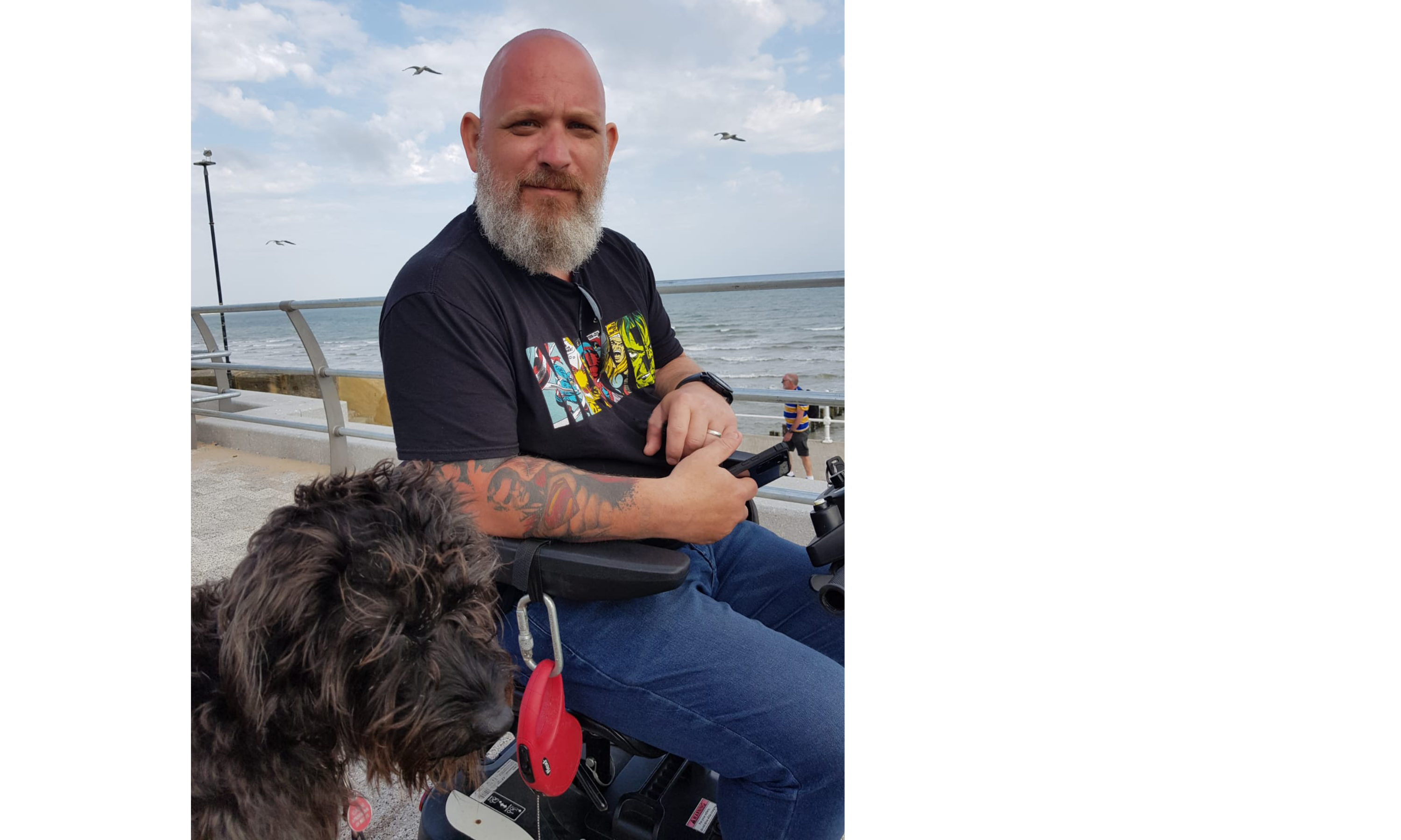 gavin camm sitting in his wheelchair at the seaside with his dog