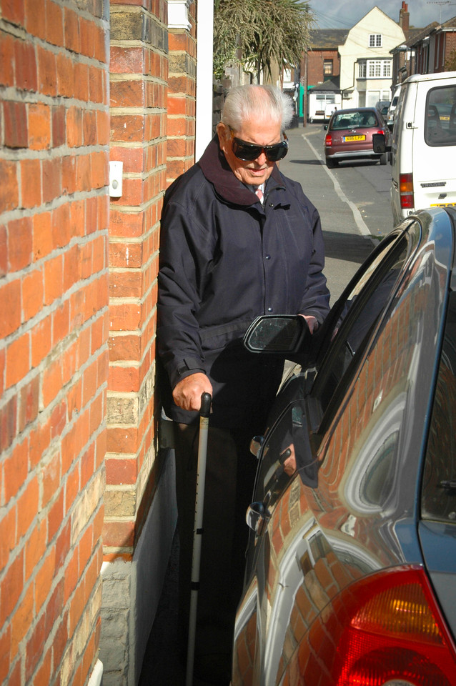 blind man trying to squeeze past a car parked on the pavement