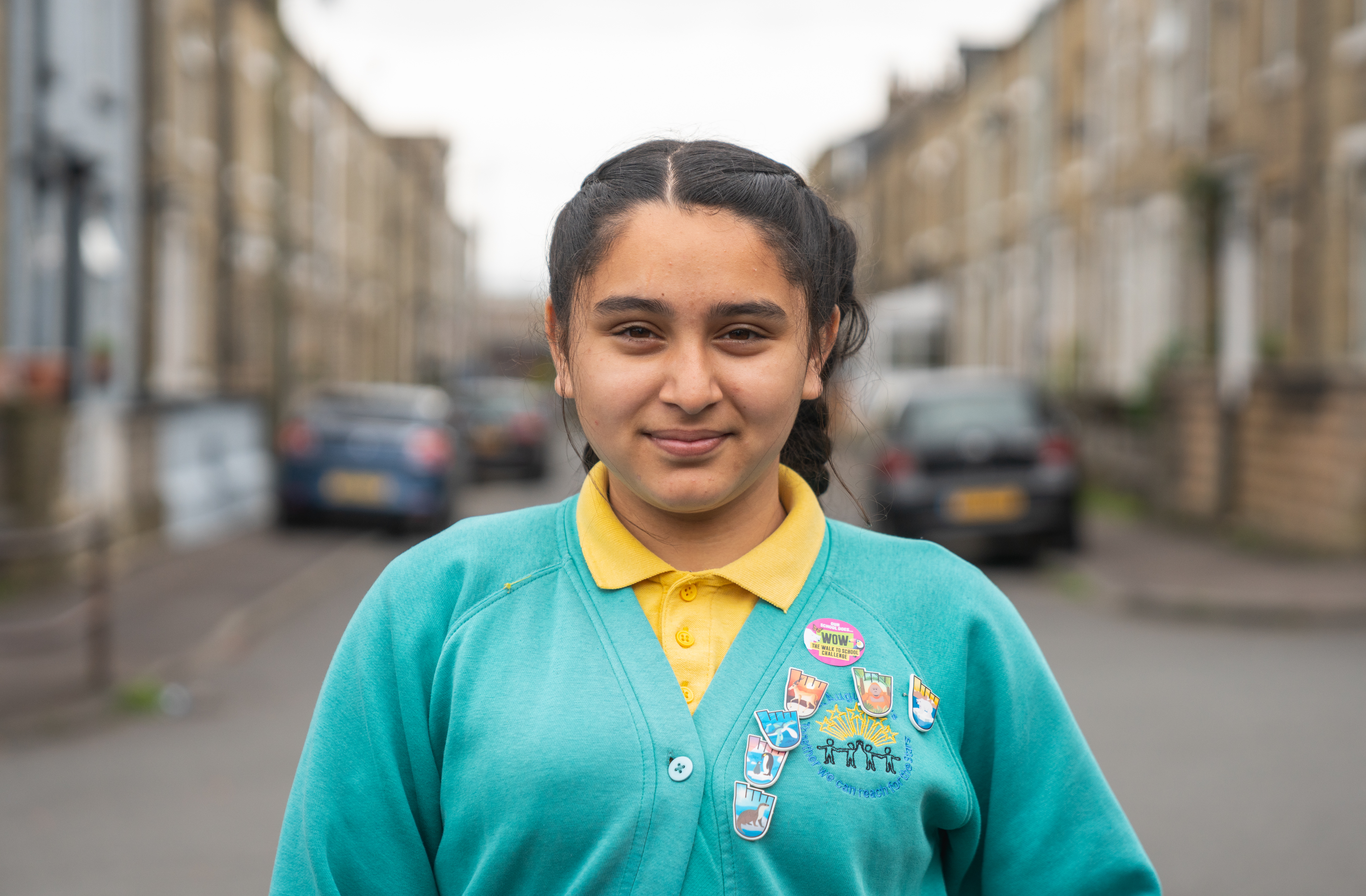 an Asian girl in school uniform wearing badges on her cardigan stands on a street and smiles