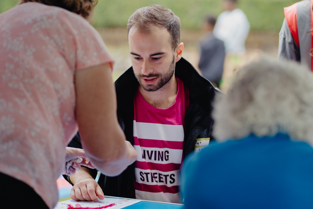 A photograph of Zak Viney at the Hanley Park event. Zak is white with brown hair and a brown beard, wearing a pink Living Streets tshirt. Zak is sat down at a stall and surrounded by other people in the foreground and background who are out of focus.