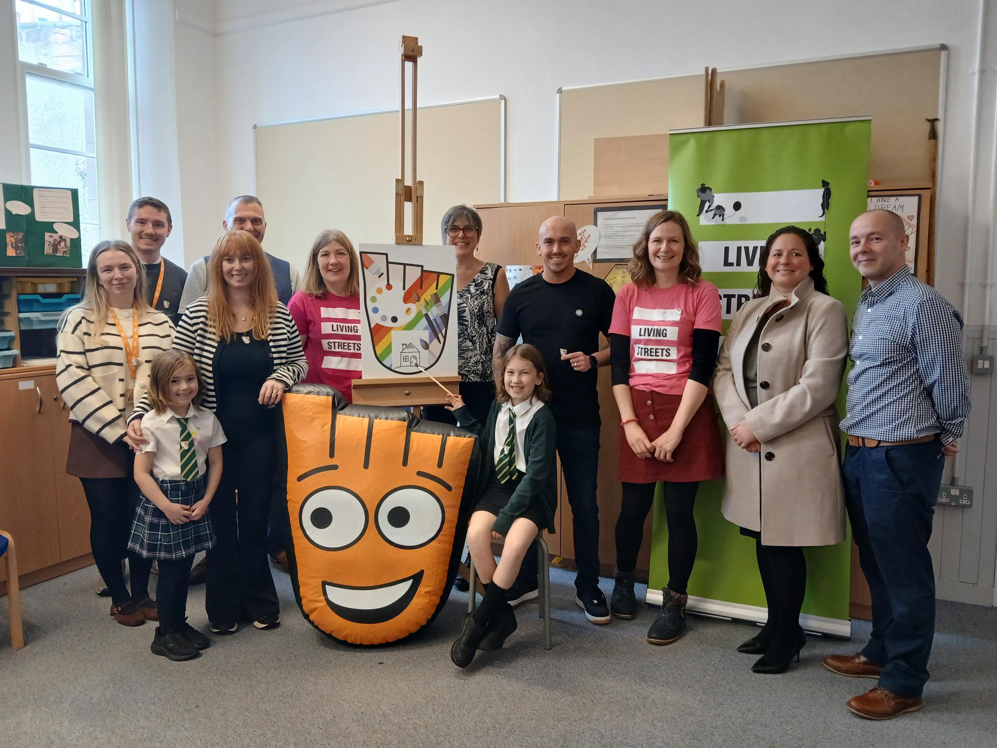 Staff, pupils and supporters pose for a group photo at Holmston Primary School in Ayr during a Living Streets Scotland award event. Two pupils stand with the school's winning artwork, alongside the Living Streets mascot, Strider, and a Living Streets Scotland banner.