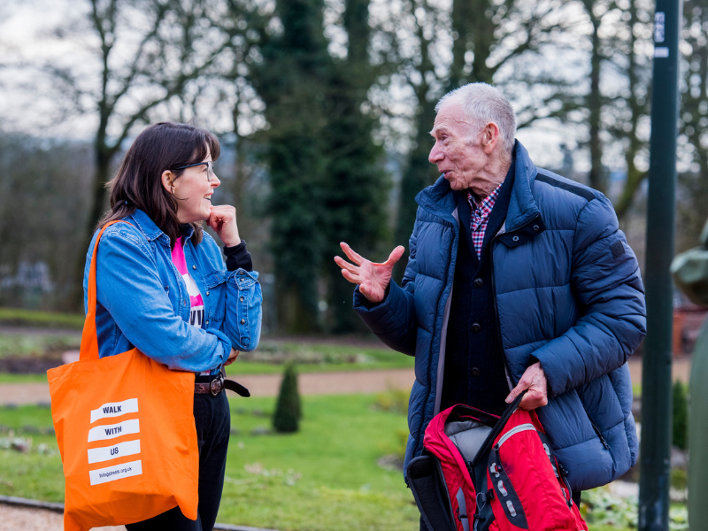 two people chatting a park one is wearing  a living streets bag