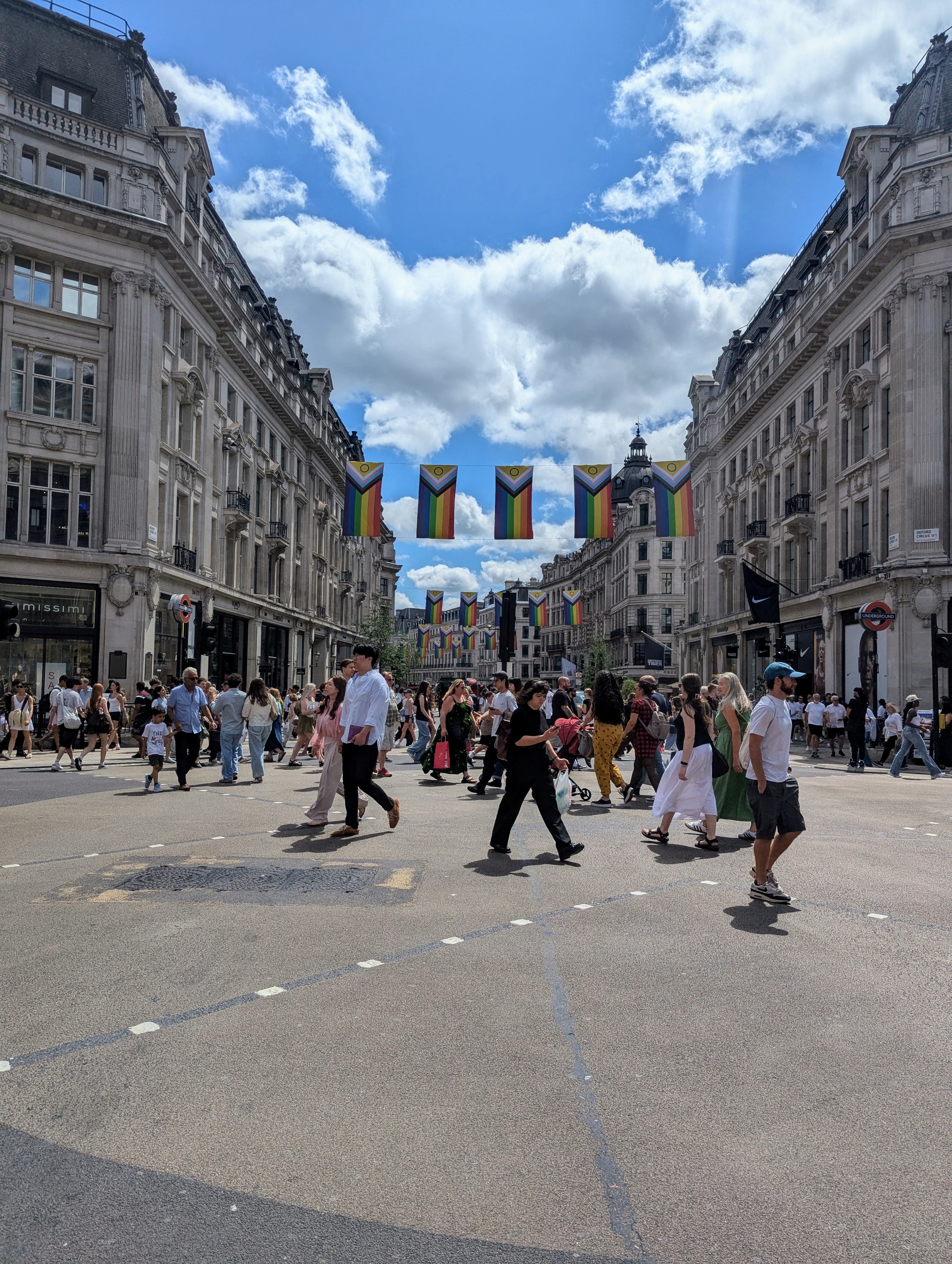 People cross a busy junction on Oxford Street in central London beneath rows of Progress Pride flags strung between historic buildings. A bright blue sky with large white clouds fills the background.