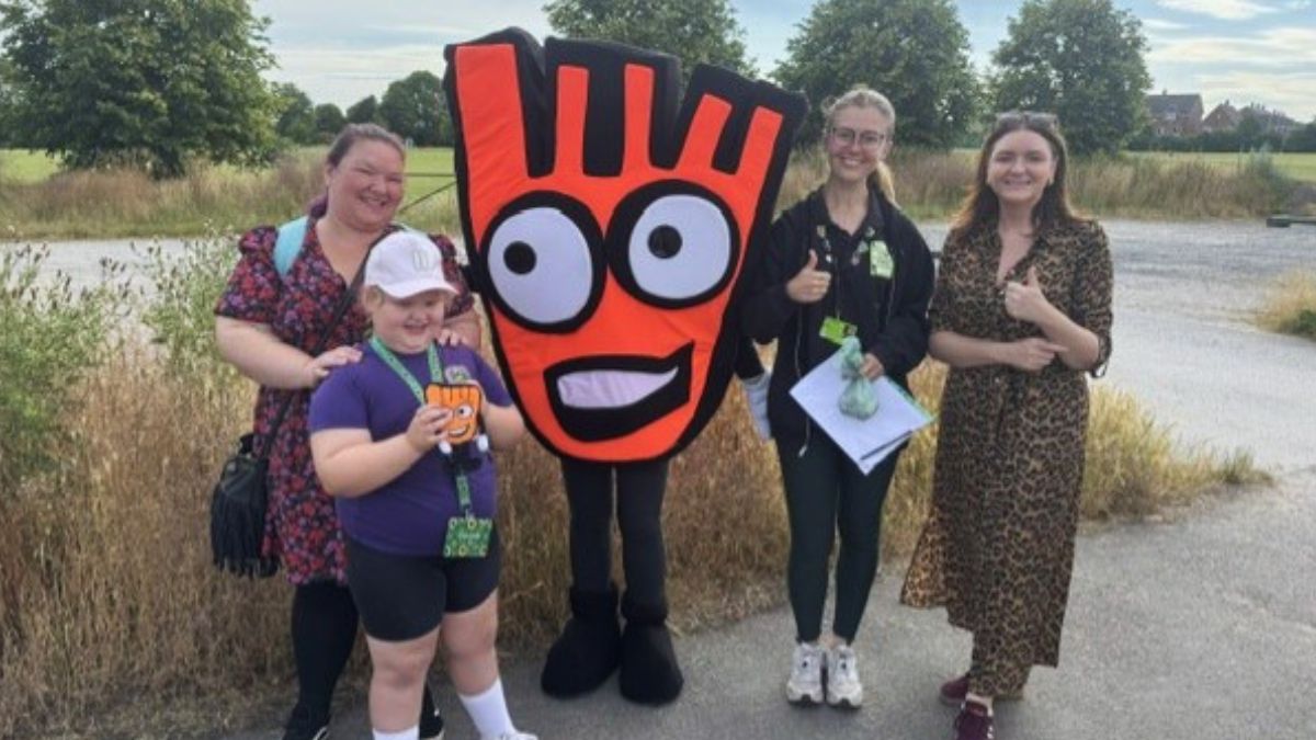 three adults and a child standing outside on  grassy path with Strider, the Living Streets mascot