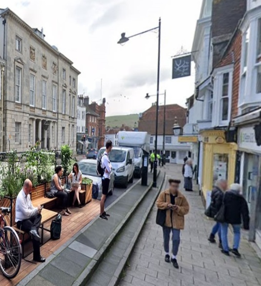 A mocked up image of potential regeneration on Lewes high street. It shows a section where car parking bays have been removed to make way for a pedestrian parklet with greenery, benches, and bicycle locking facilities.
