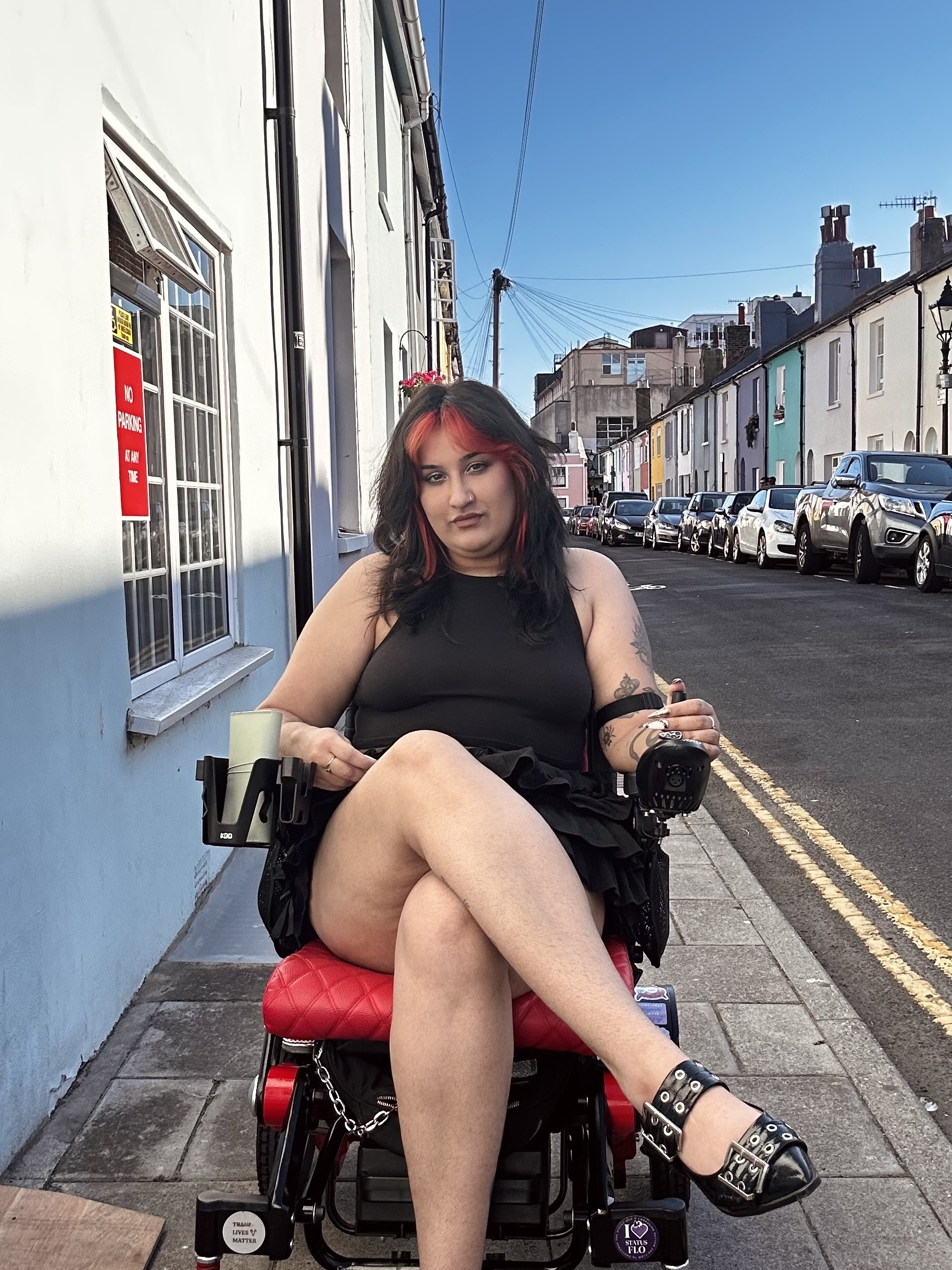 A person sits in a powered wheelchair on a narrow pavement along a residential street. The wheelchair has a red seat and visible joystick controls. The person is wearing a sleeveless black top, a black skirt and black buckle shoes, with black hair featuring red highlights. A row of colourful terraced houses and parked cars lines the street behind them under a clear blue sky. A red sign in a nearby window reads “NO PARKING AT ANY TIME.”