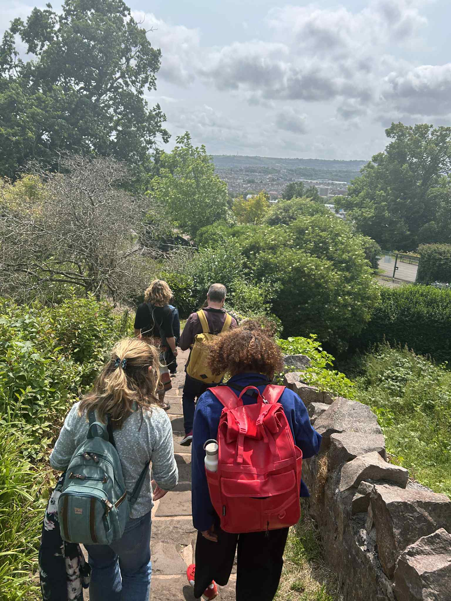 A photograph of four people pictured from behind. The people are in the foregound talking to one another and walking down a set of stone steps. In the background are tree tops and a view over the Bristol city skyline.