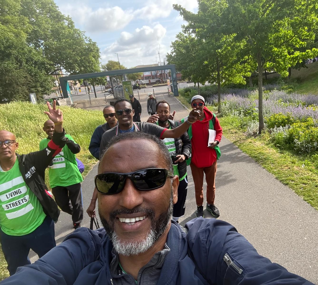Seven members of Living Streets South London Somali Men's Group take a selfie of themselves as they walk close together down a sunny pathway through a park. They are smiling and raising their hands. On both sides of the path the sun shines down on grassy beds and wildflowers.