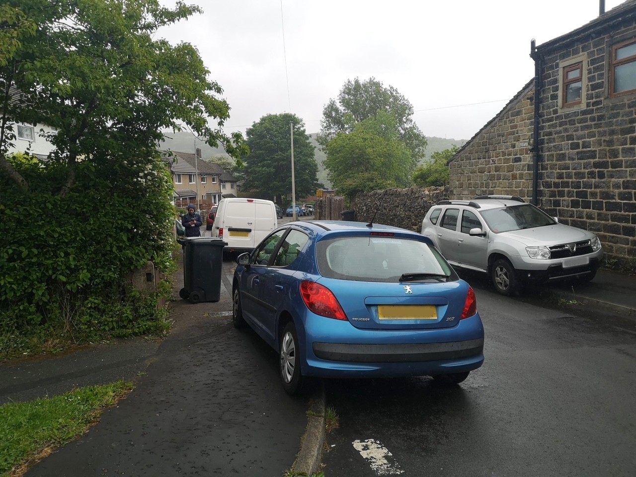 a blue car and a silver car are parked on the pavement besides a wheelie bin, causing an obstruction to the path