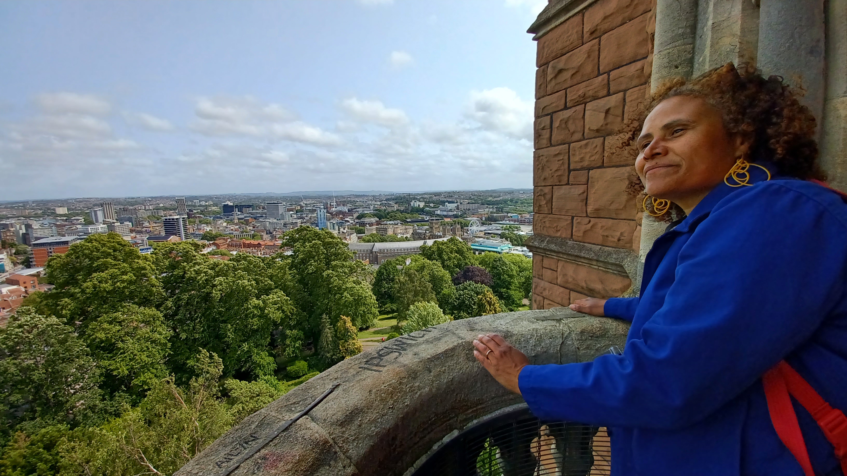 A photograph of a woman standing on a stone balcony looking at the view below, which is sunny tree tops and the Bristol city skyline.