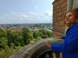 A photograph of a woman standing on a stone balcony looking at the view below, which is sunny tree tops and the Bristol city skyline.
