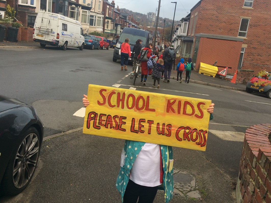 A child standing on a residential street holding a placard that reads School Kids: Please Let Us Cross