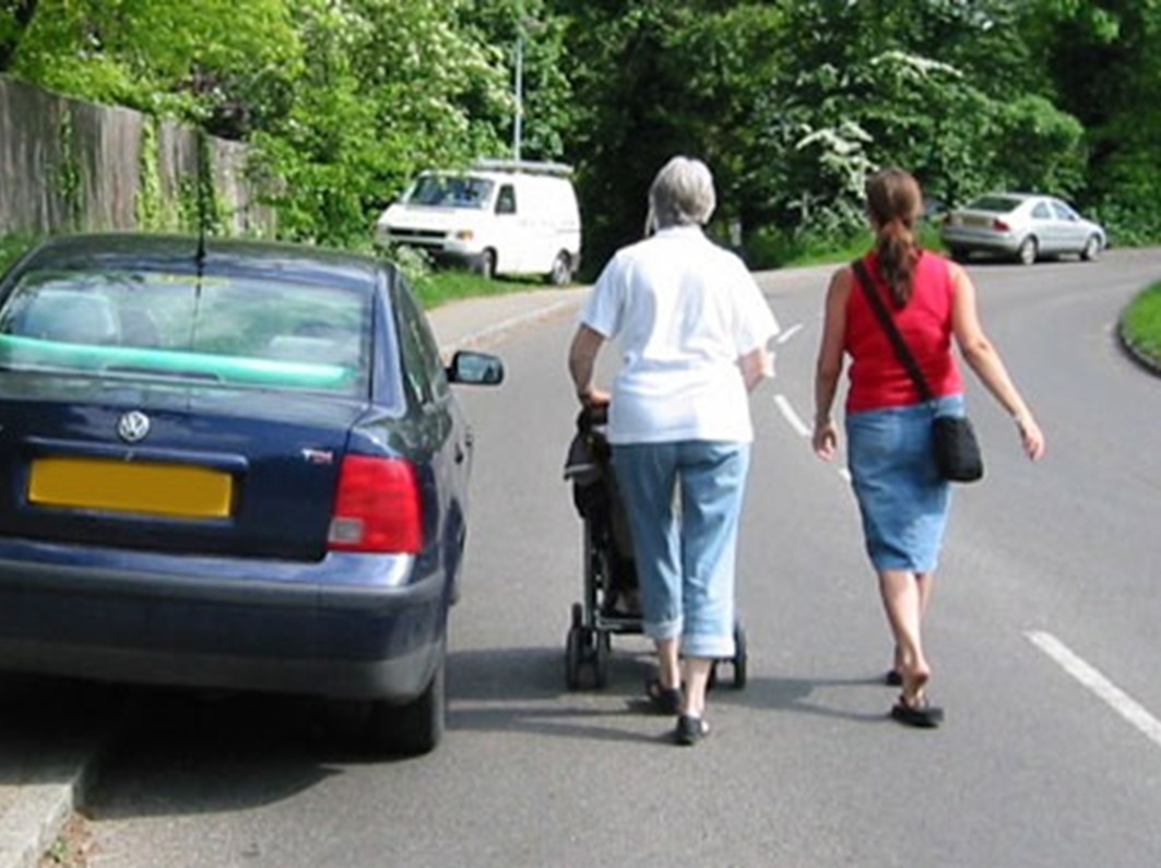 Two women (one pushing a pushchair) walk in the road because a car is blocking the pavement