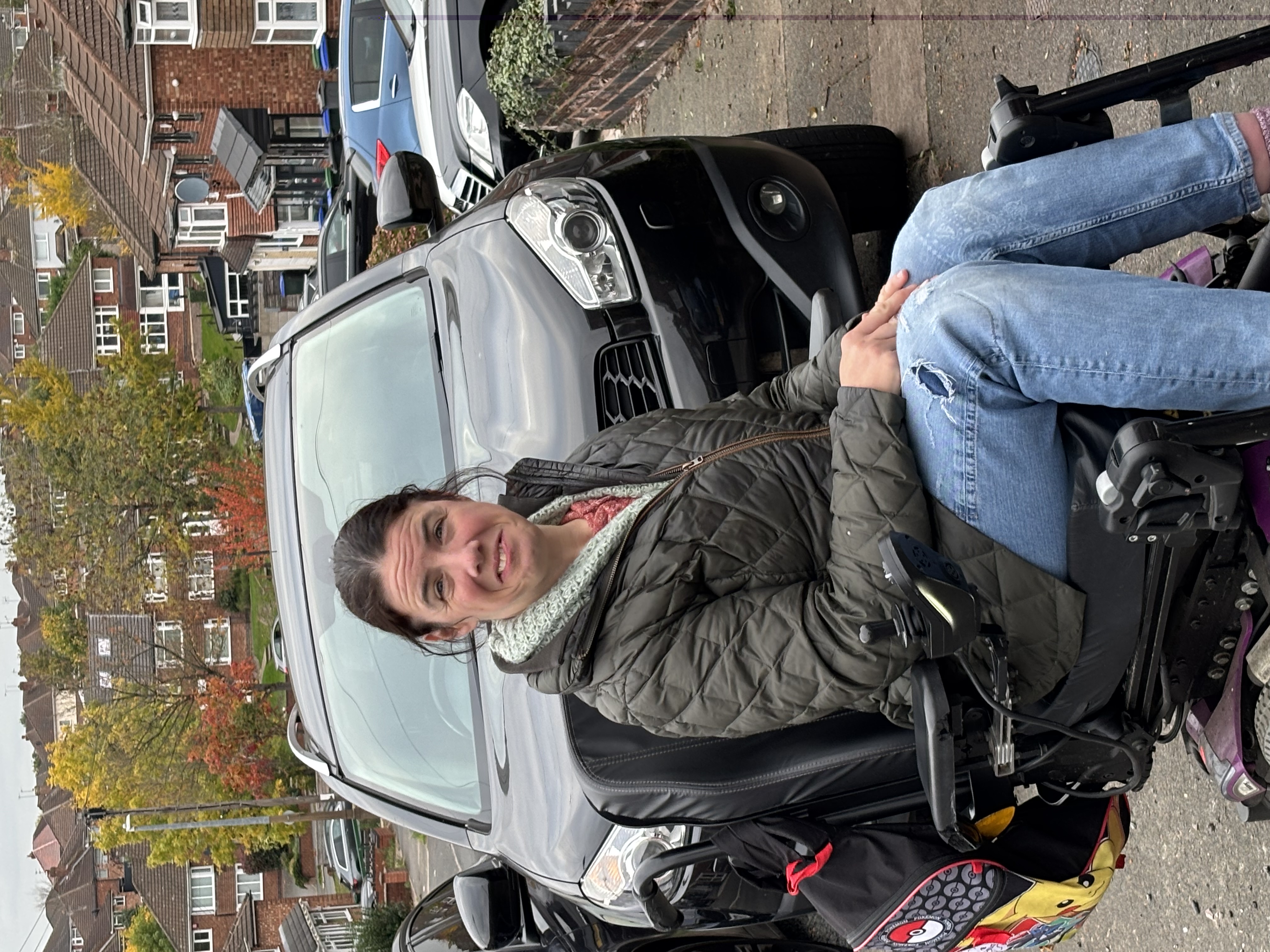 Aideen poses in her power wheelchair in front of a pavement parked car
