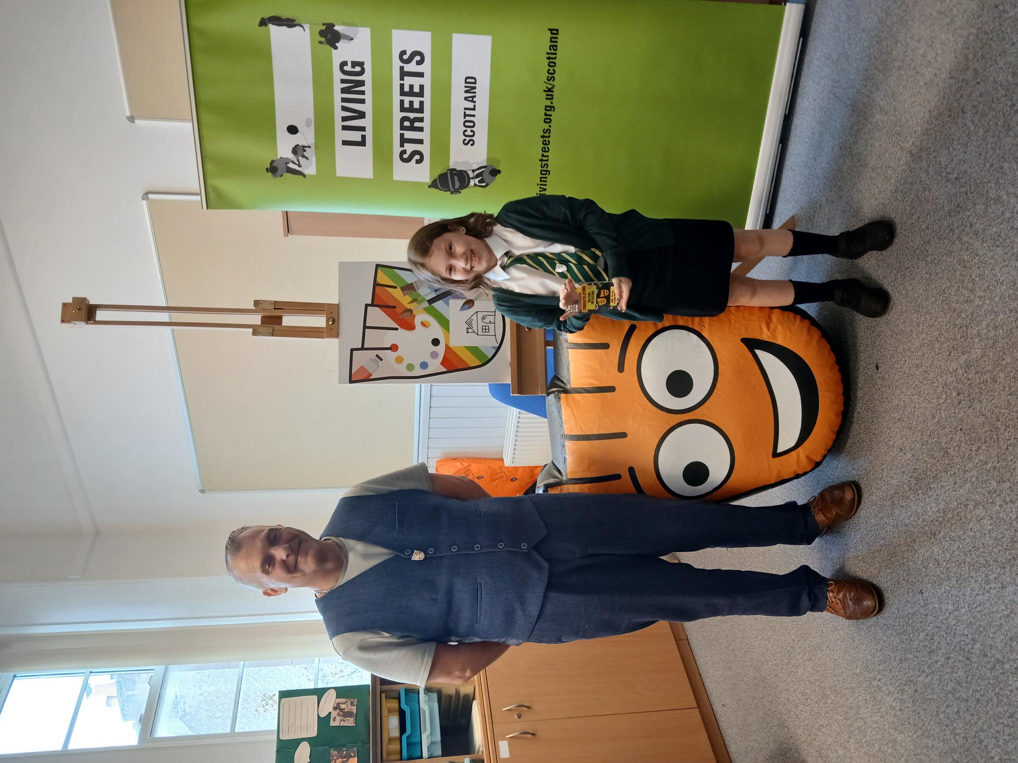 A pupil in school uniform holds a Walking Hero award certificate while standing beside an adult in a classroom. A Living Streets Scotland banner and the charity's orange mascot are visible in the background during an award presentation at Holmston Primary School in Ayr.