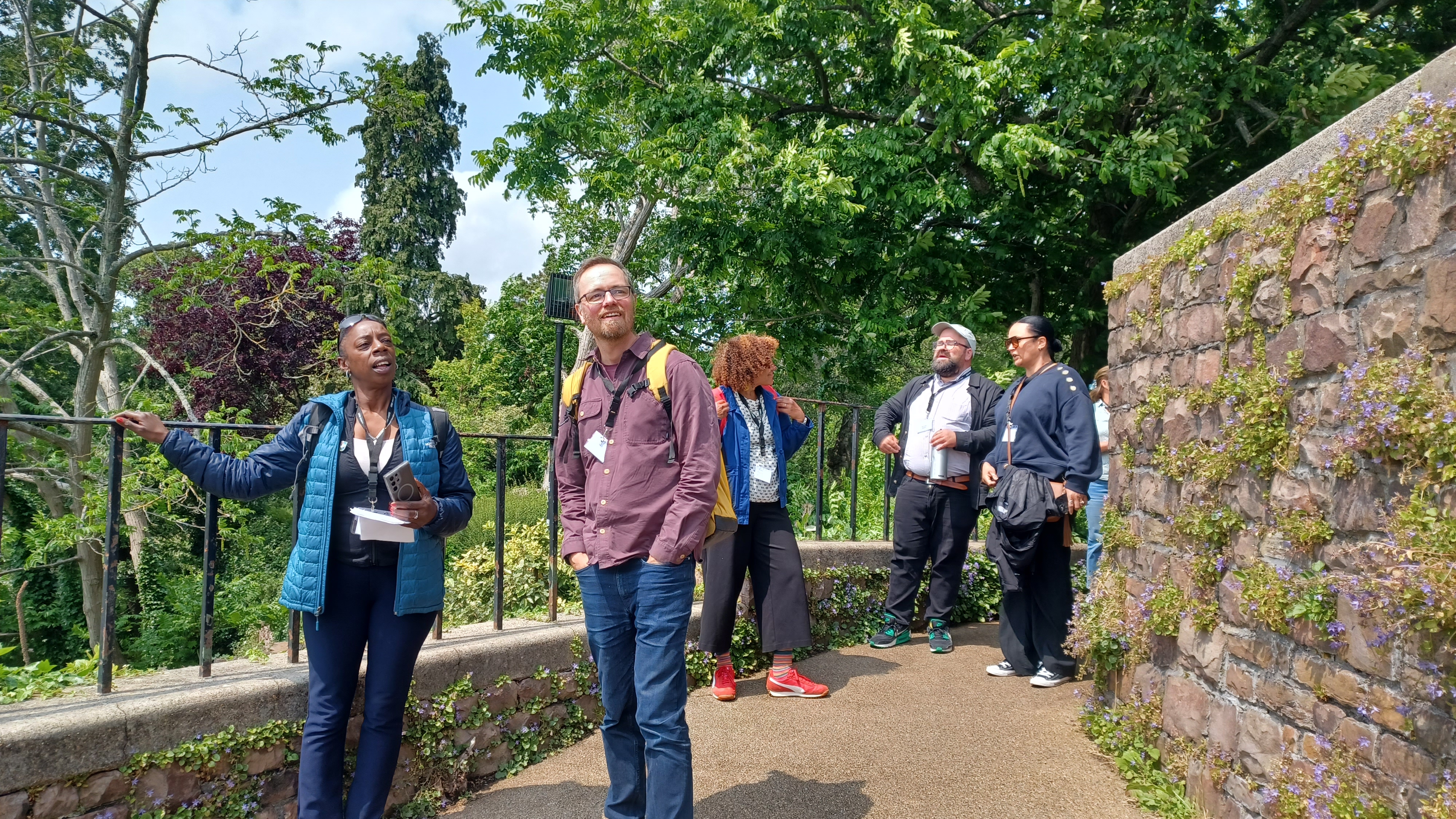 A photograph of five people. They are loosely spread out and talking to one another as they walk along. In the background are green trees and bushes, and on the right hand side of the image is a stone wall with purple flowers growing through the cracks between the stones.