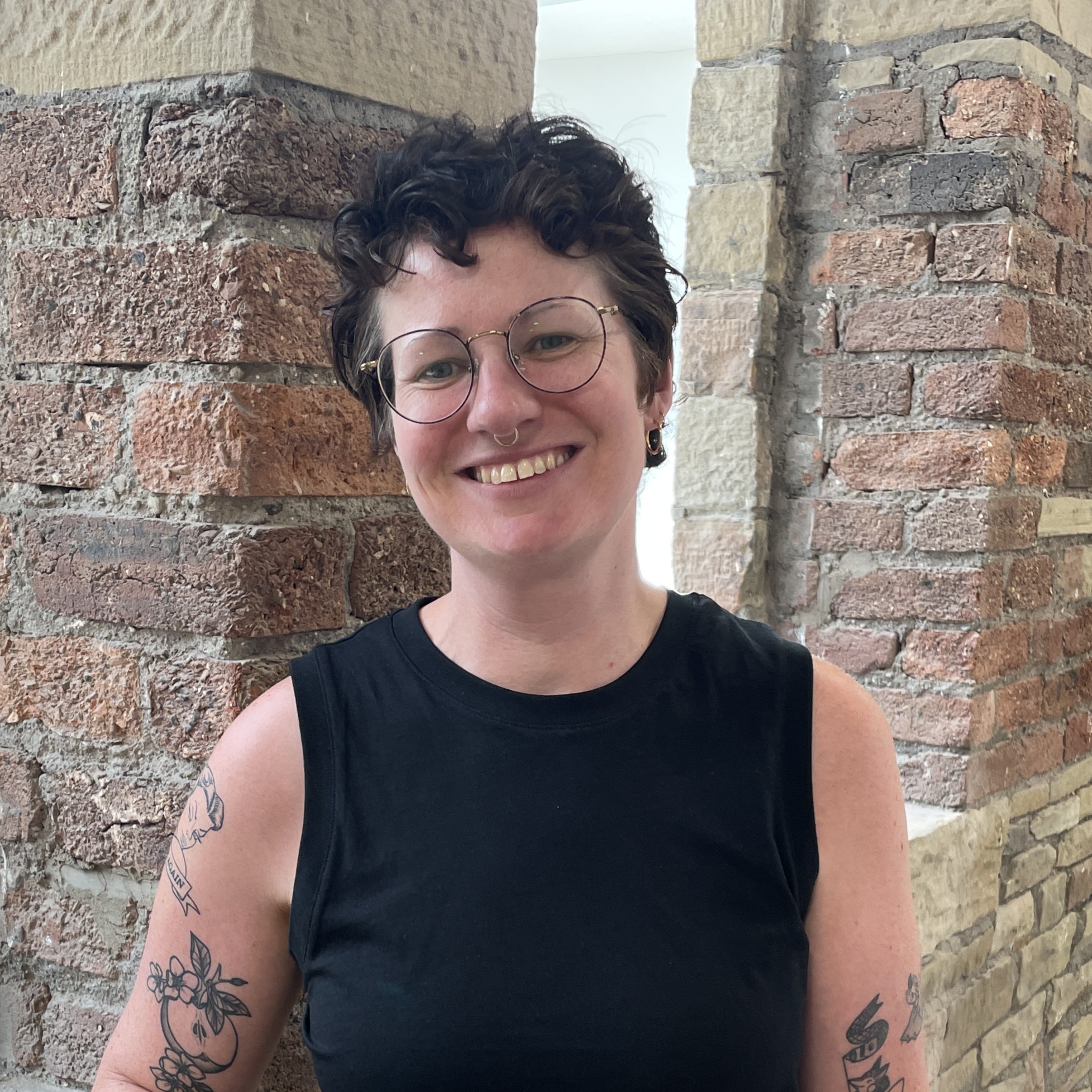 An image of Hannah Davies, a white person with short brown hair wearing glasses and smiling at the camera. There is a brick wall and windows in the background.