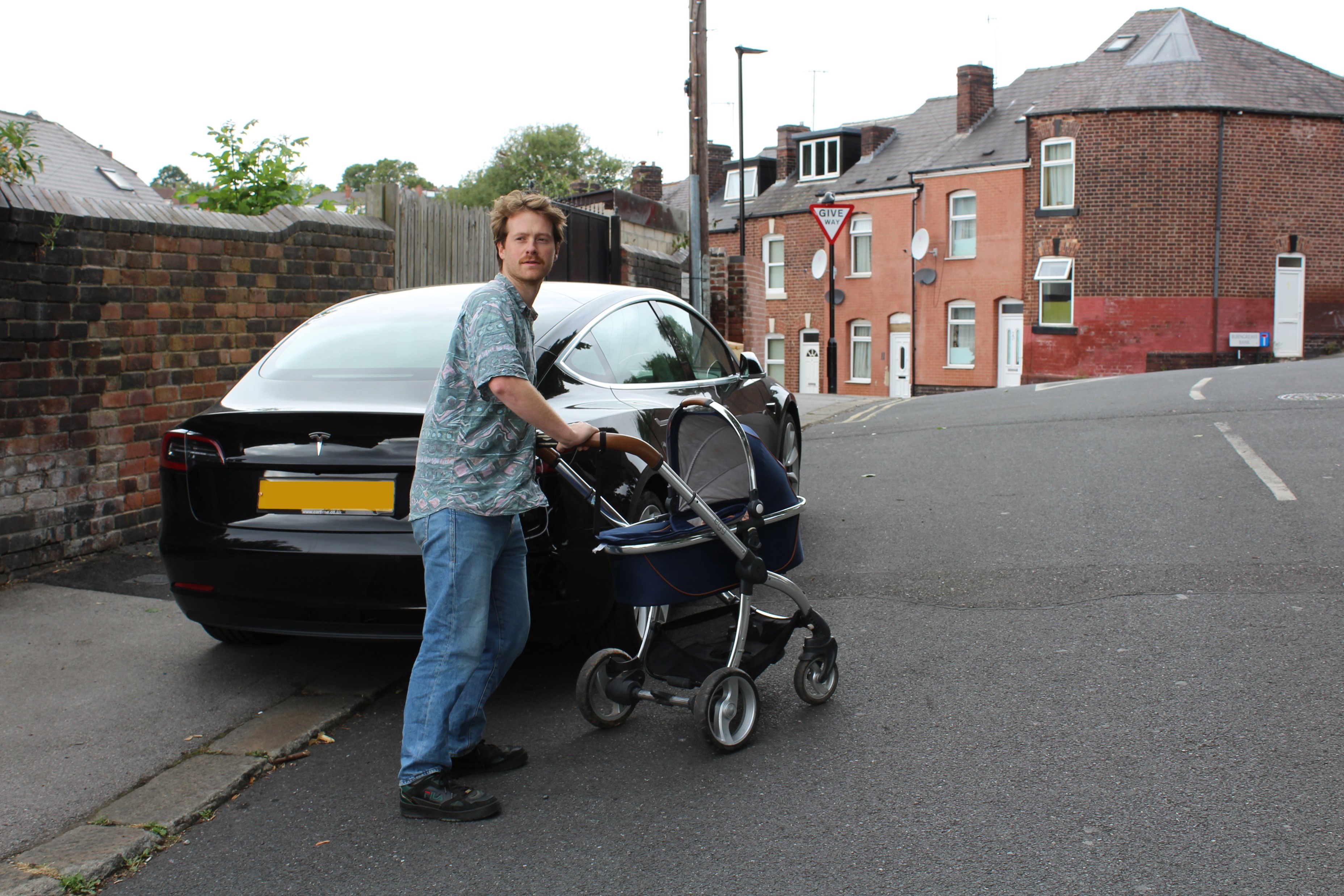 A parent moves out into the road pushing a pram to avoid a pavement parked car