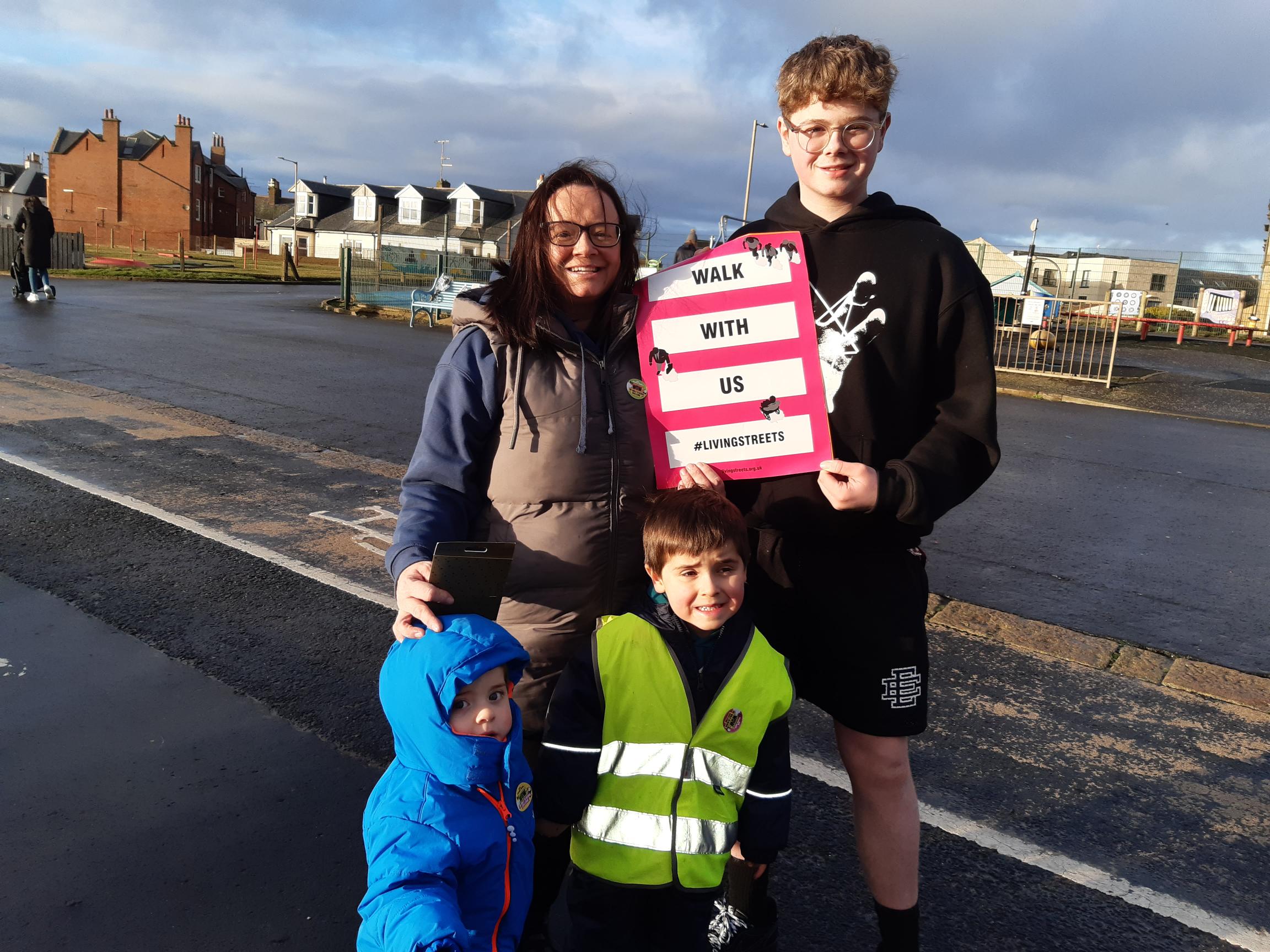 A mother and her three children smile at the camera. The eldest boy holds up a 'Walk with us' Living Streets poster.